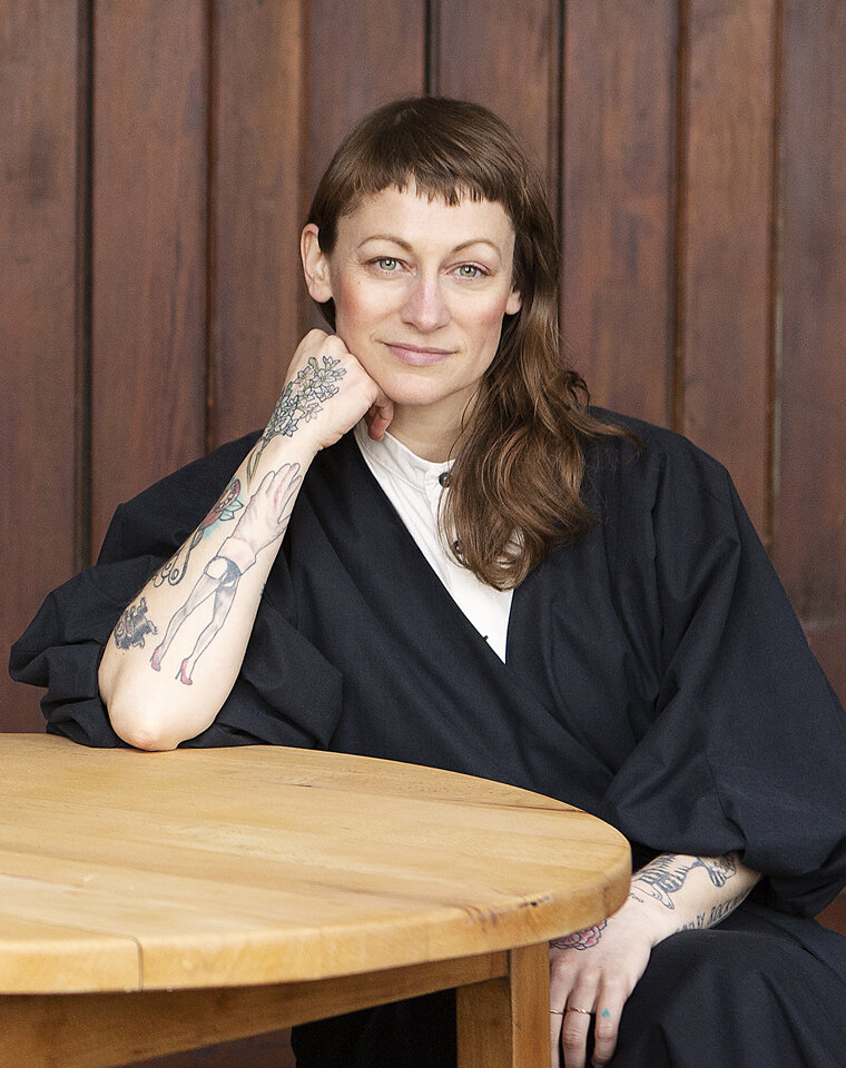 Porträt einer Frau. Sie sitzt an einem runden Tisch, im Hintergrund Holzvertäfelung.