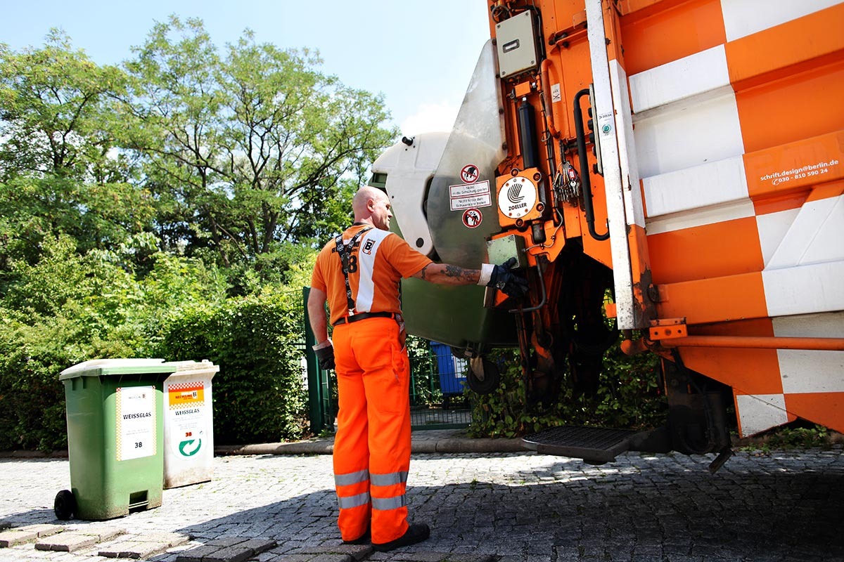 Ein Müllfahrer in oranger Arbeitskleidung entleert Mülltonnen in ein Müllfahrzeug.
