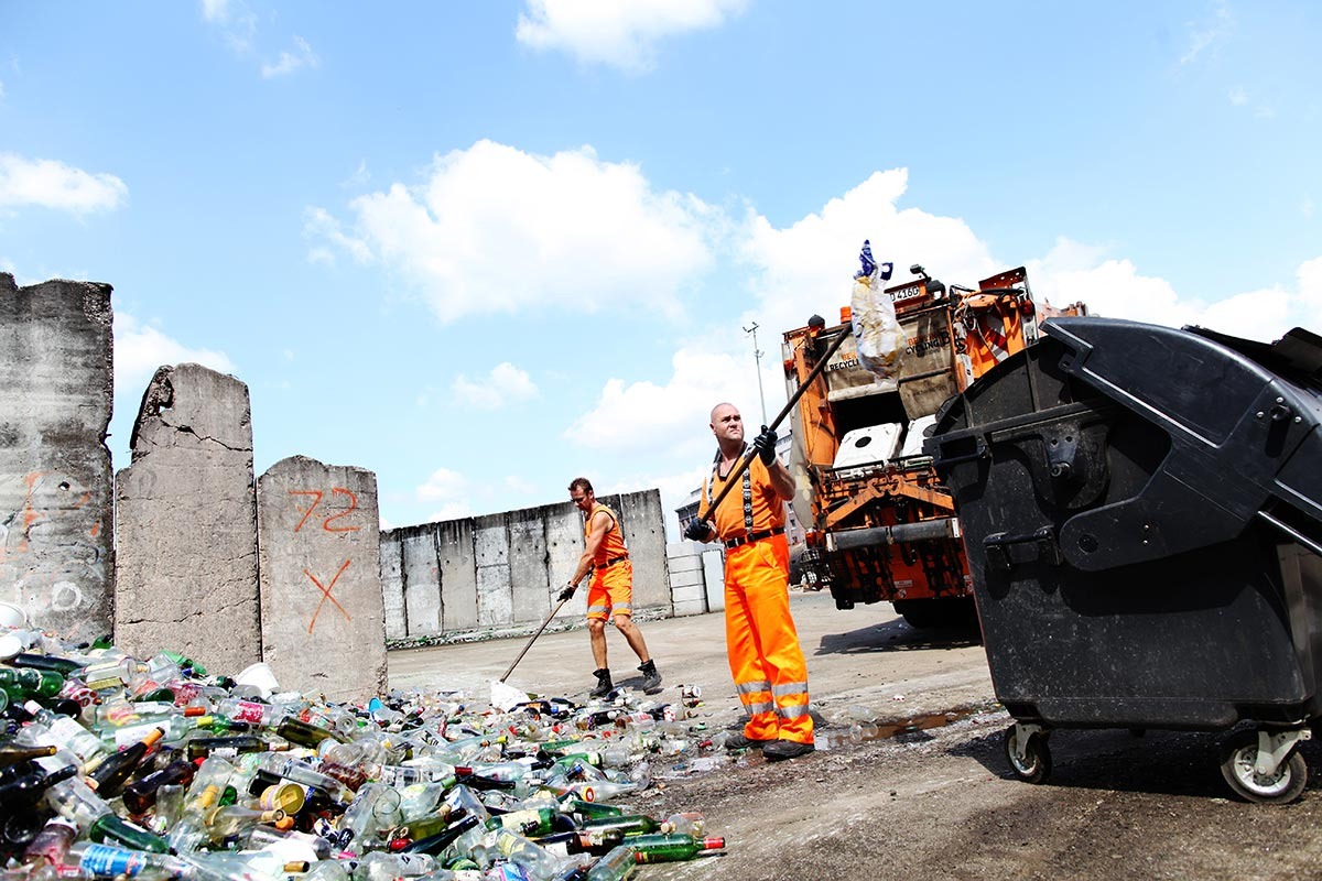 Zwei Müllmänner in oranger Arbeitskleidung sortieren Plastikmüll aus einem Haufen aus zerbrochenen Glasflaschen.
