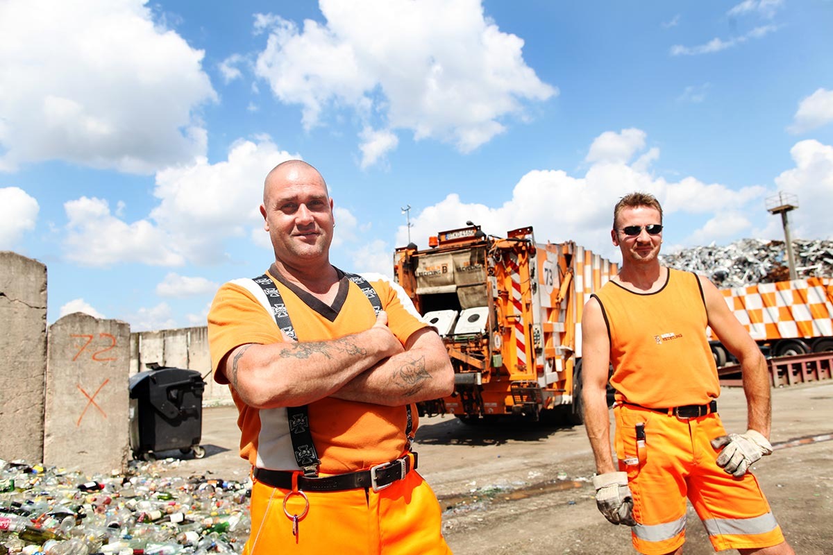 Zwei Müllmänner in oranger Arbeitskleidung posieren vor ihrem Müllfahrzeug auf einer Glassortieranlage.