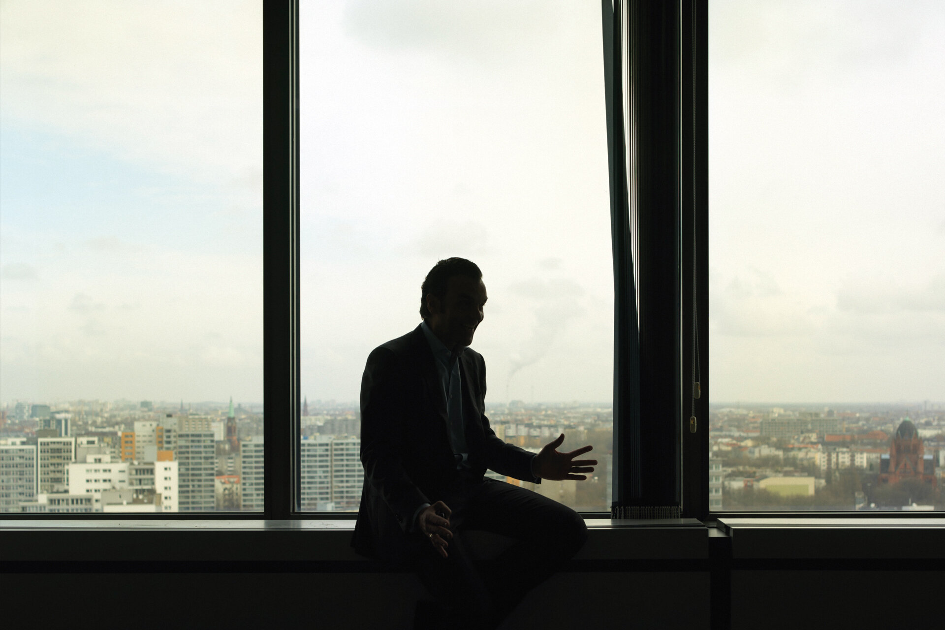 Mensch sitzt im Gegenlicht vor dem Fenster eines Hochhauses. Im Hintergrund der diesige Blick über die Häuser Berlins.