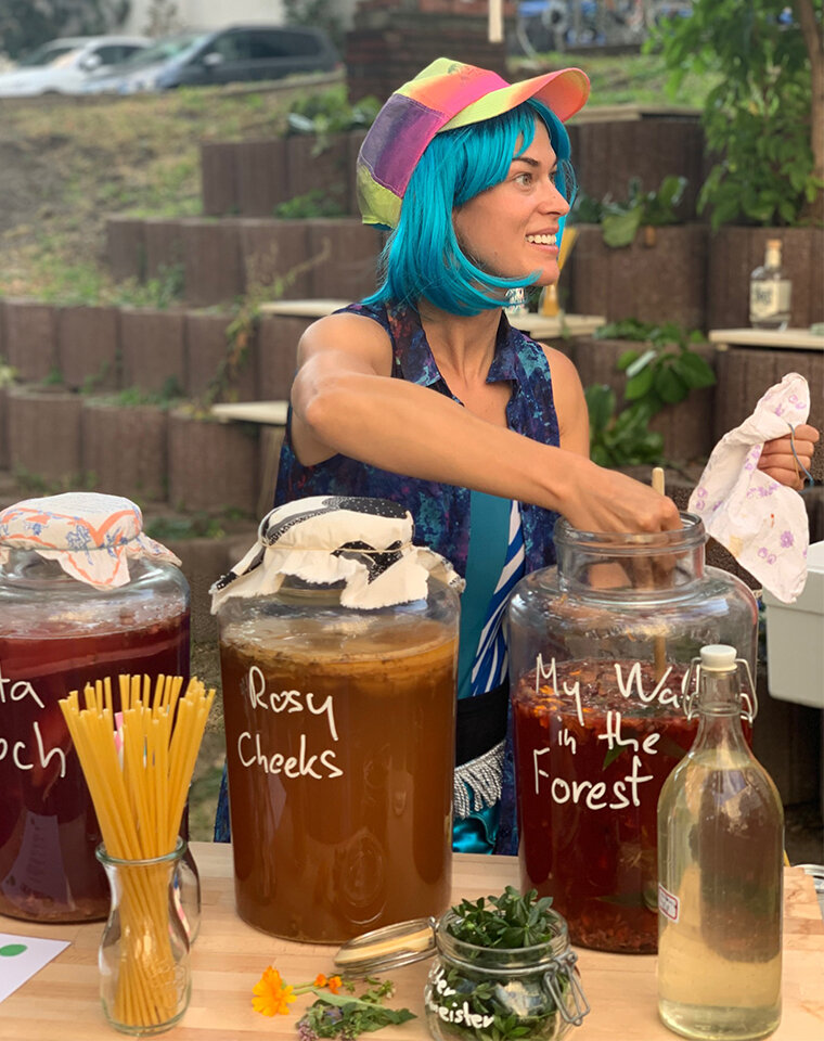 Frau mit blau gefärbten Haaren. Vor ihr stehen drei große Gläser mit fermentierten Lebensmitteln. Sie greift mit der Hand in eins der Gläser.