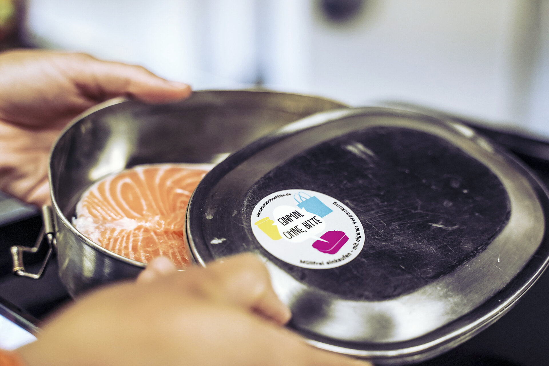 Nahaufnahme: Hände öffnen Mehrwegbox (gefüllt mit frischem Lachs). Auf der Box klebt ein Aufkleber der Initiative: Einmal ohne Bitte.