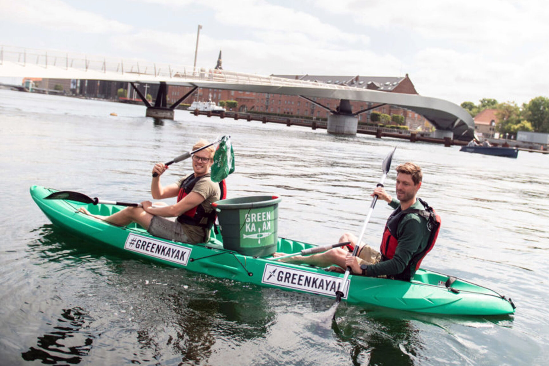 Zwei Männer paddeln auf der Spree in einem grünen Kayak und sammeln Müll aus dem Wasser.