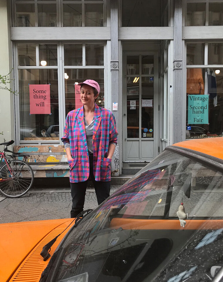 Eine Frau steht in einem bunt karierten Jackett und rosa Cap vor einem Secondhandladen.