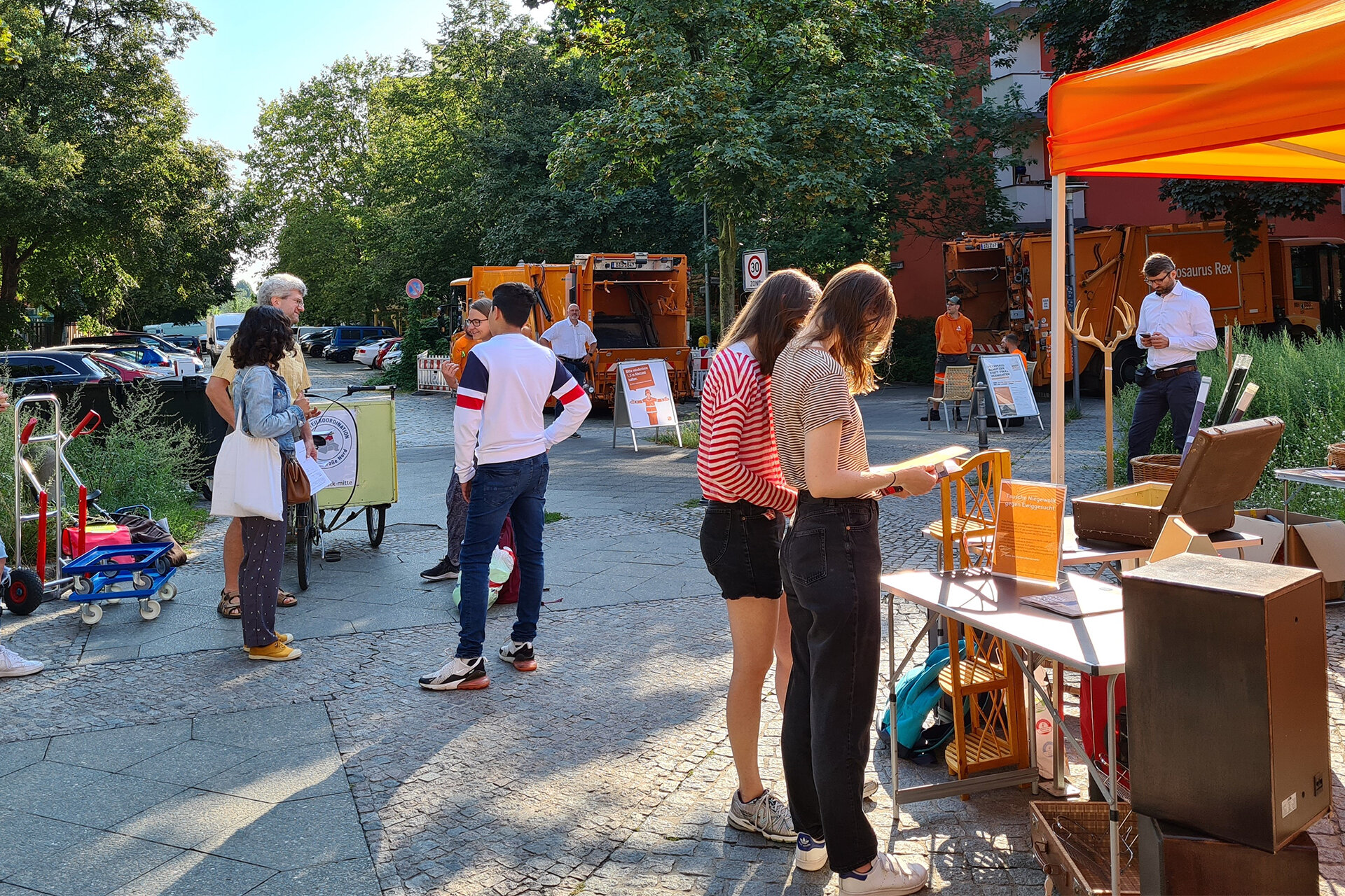 Verschiedene Menschen stehen auf der Straße und reden miteinander. Überall steht gut Erhaltenes zum Weitergeben. Zwei Frauen stehen im Vordergrund an einem Infostand und schauen sich Infomaterial der BSR an. Im Hintergrund zwei Müllwagen und BSR-Mitarbeiter.