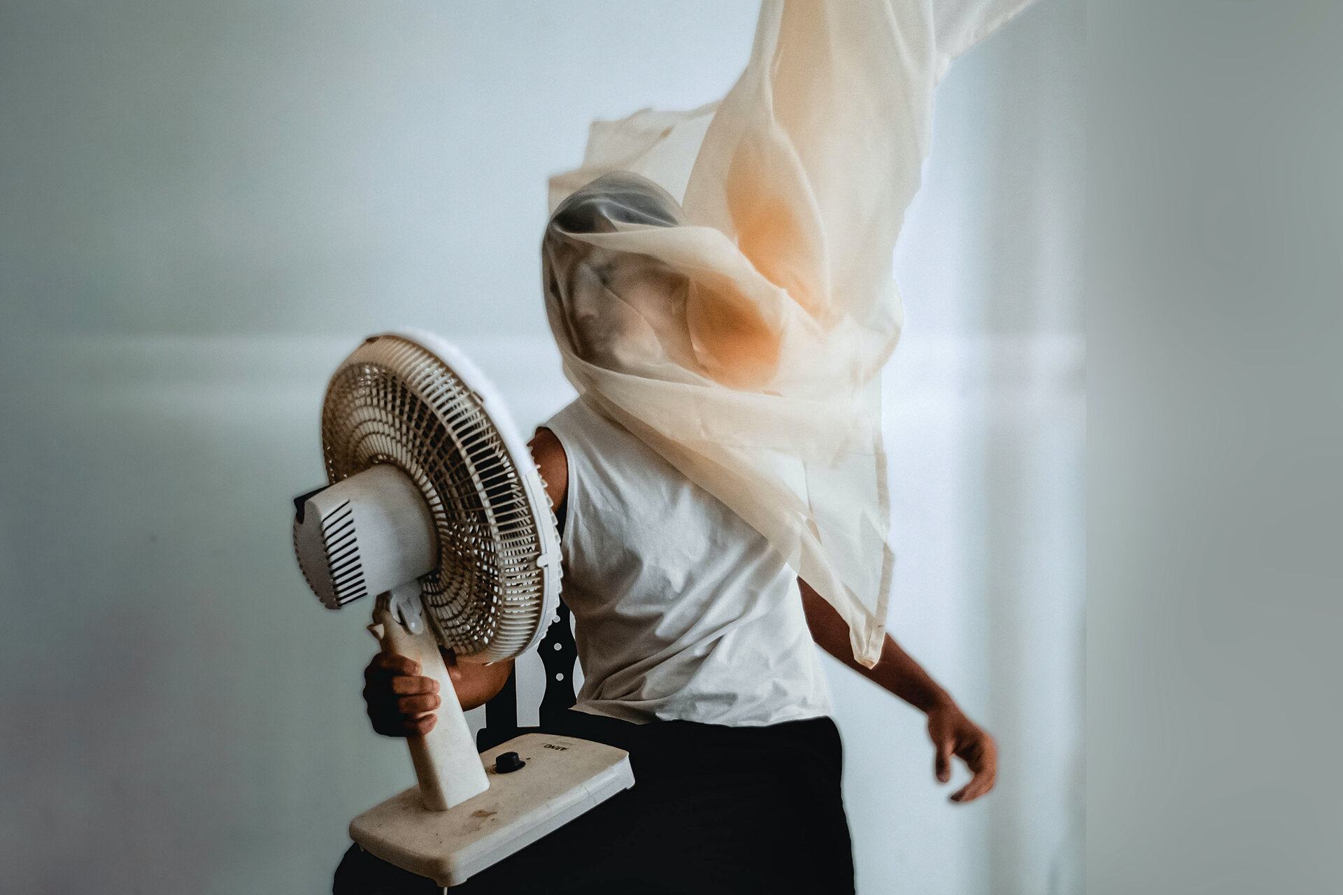 Mann mit Tuch über dem Kopf hält Ventilator in der Hand und pustet sich Luft zu. Das Tuch weht im Wind.