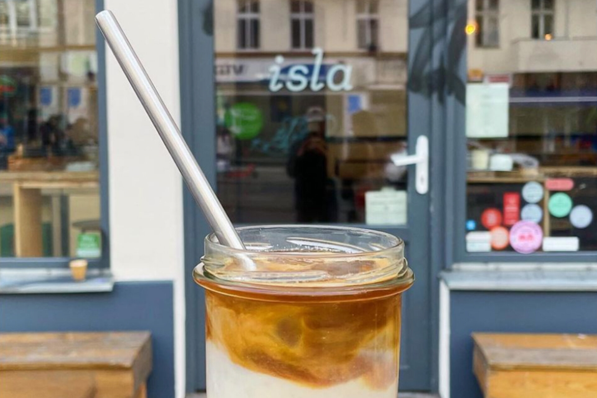 Eine Frauenhand hält ein Einweckglas mit heller und brauner Flüssigkeit vor der Eingangstür von Isla Coffee.