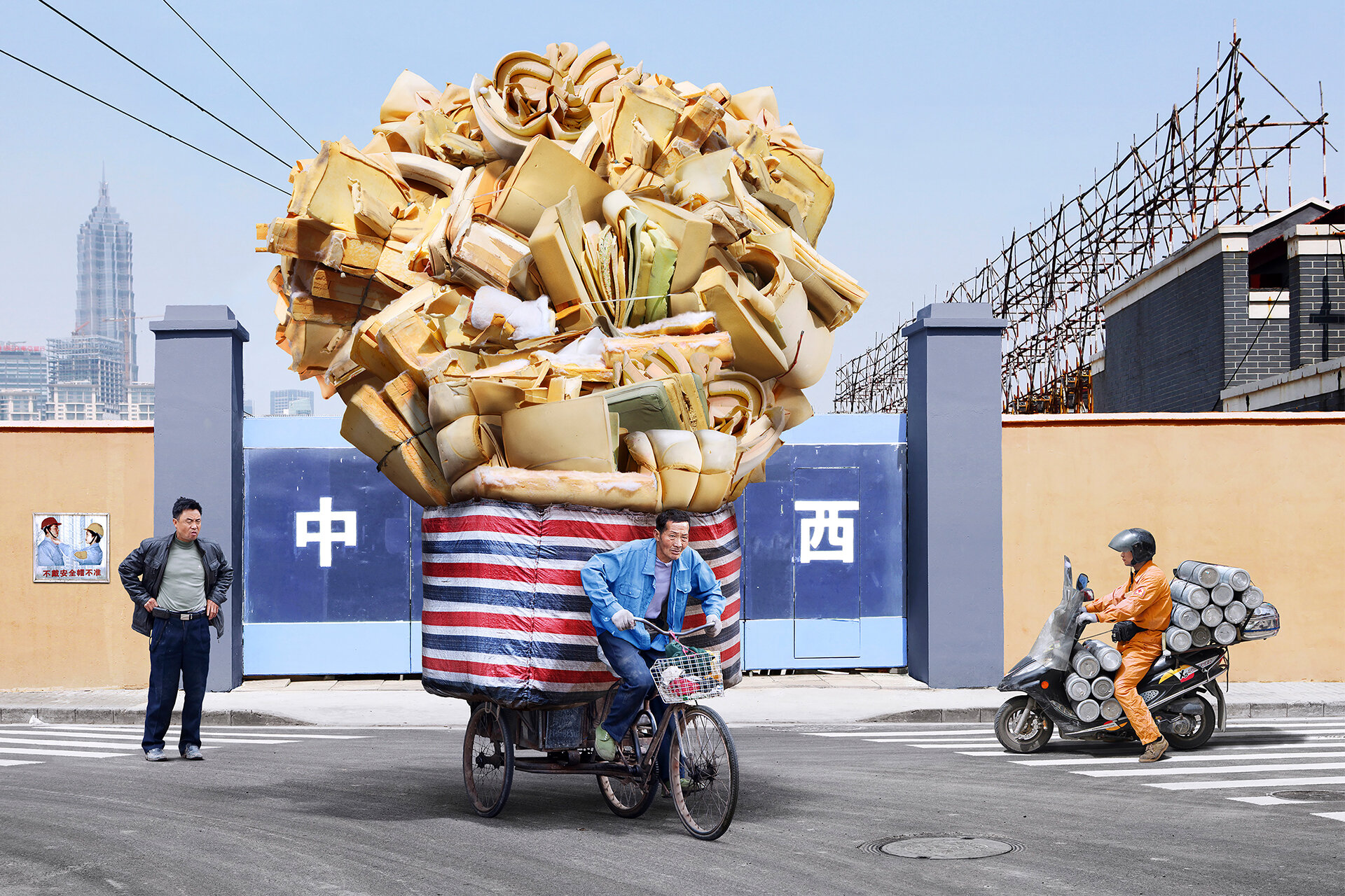 Vor einer Mauer tritt ein Asiate auf seinem Fahrrad kräftig in die Pedale. Das Fahrrad ist beladen mit übermäßig vielen Schaustoffen. Ein anderer Mann steht staunend daneben. Ein anderer Mann in Orange auf seinem mit grauen Rollen beladenen Moped lässt das Fahrrad passieren. Hinter der Mauer sind hohe Gebäude und ein im Bau befindliches Haus sichtbar.