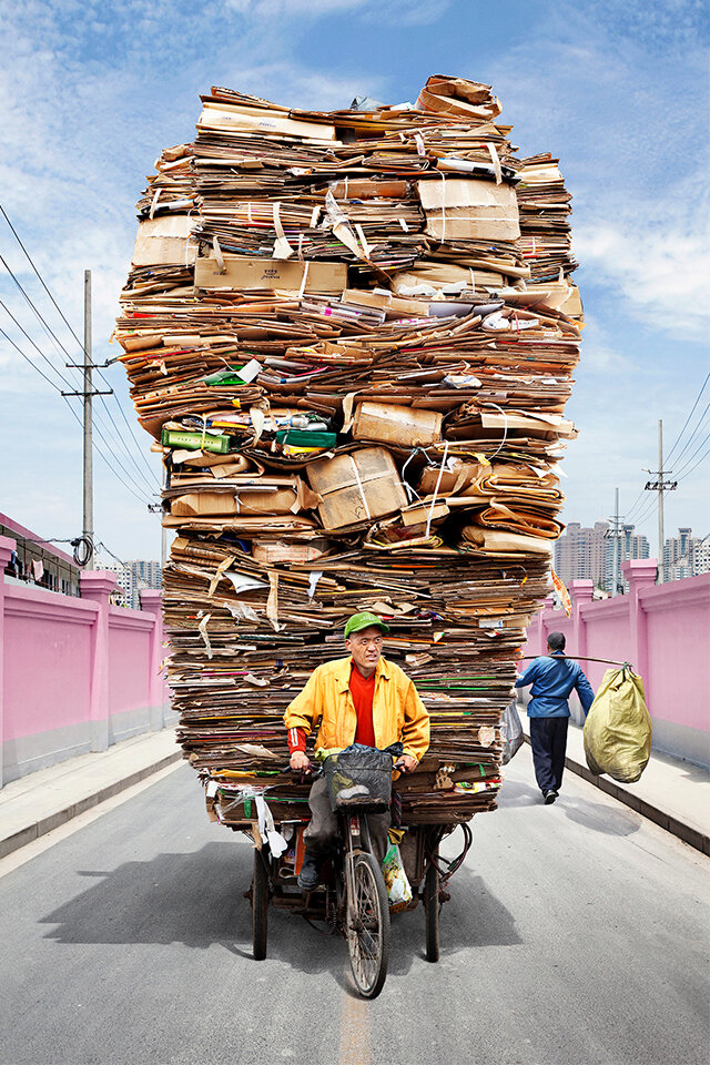 Flankiert von einer pinken Mauer tritt ein Asiate auf seinem Fahrrad kräftig in die Pedale. Das Fahrrad ist beladen mit kiloweise ordentlich gefalteten und gestapelten Pappkartons. Ein anderer Mann läuft im Hintergrund vorbei und balanciert zwei an einer Stange befestigten Säcke auf seiner Schulter. Im Hintergrund eine Stadtkulisse.