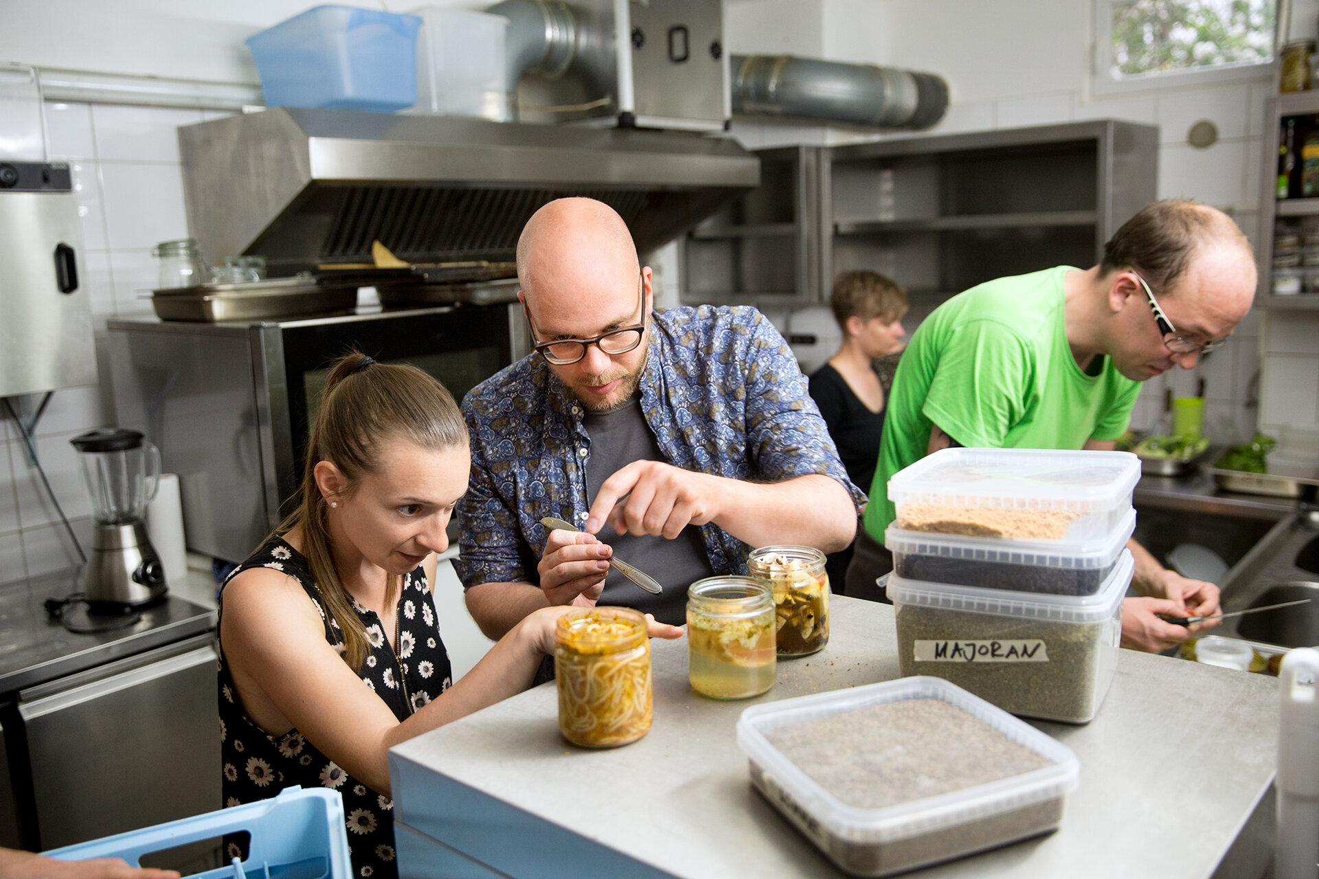 In einer Küche versuchen sich mehrere Workshop-Teilnehmer und Teilnehmerinnen am Fermentationsprozess. Eine Frau und ein Mann deuten dabei auf eines von drei mit Lebensmitteln gefüllten Einweckgläsern.