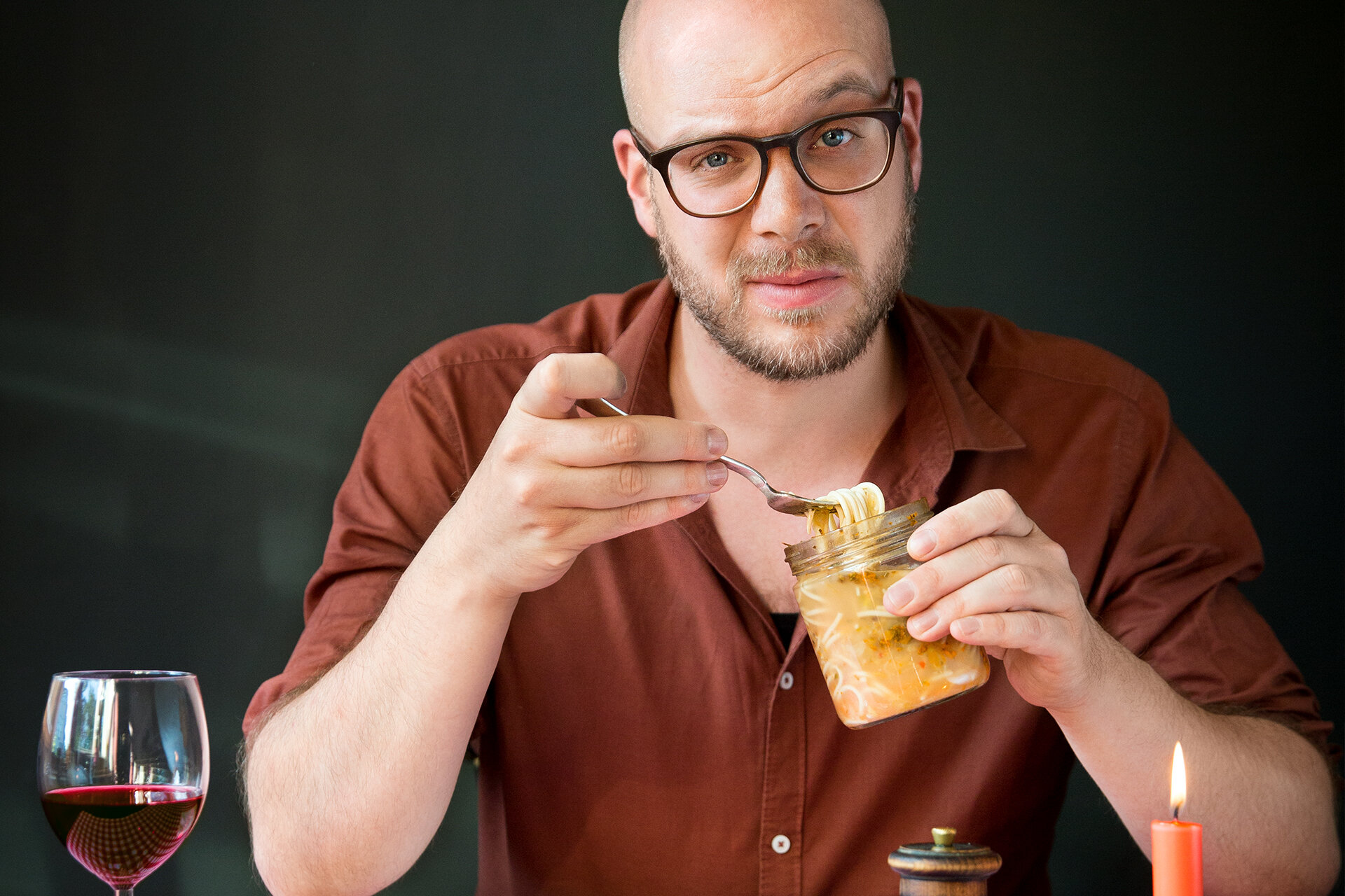 Ein am Tisch sitzender Mann schaut mit hochgezogener Augenbraue in die Kamera. In der Hand hält er ein Einweckglas mit fermentierten Spaghetti.