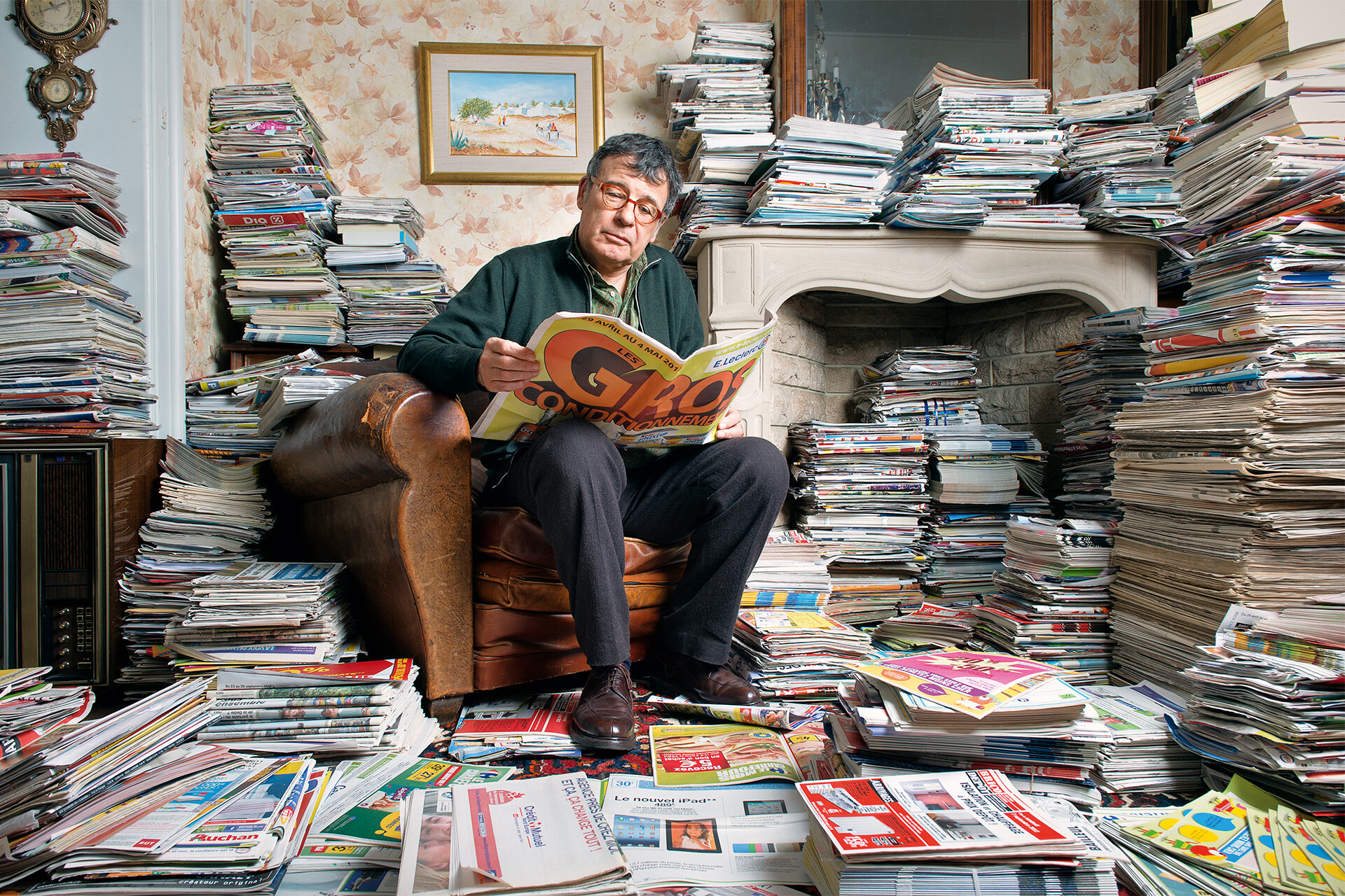 In seinem Wohnzimmer mit Kamin voller tausender ordentlich gestapelter Zeitschriften, Magazinen und Zeitungen, sitz ein Mann in seinem Sessel und liest lächelnd eine Zeitung.