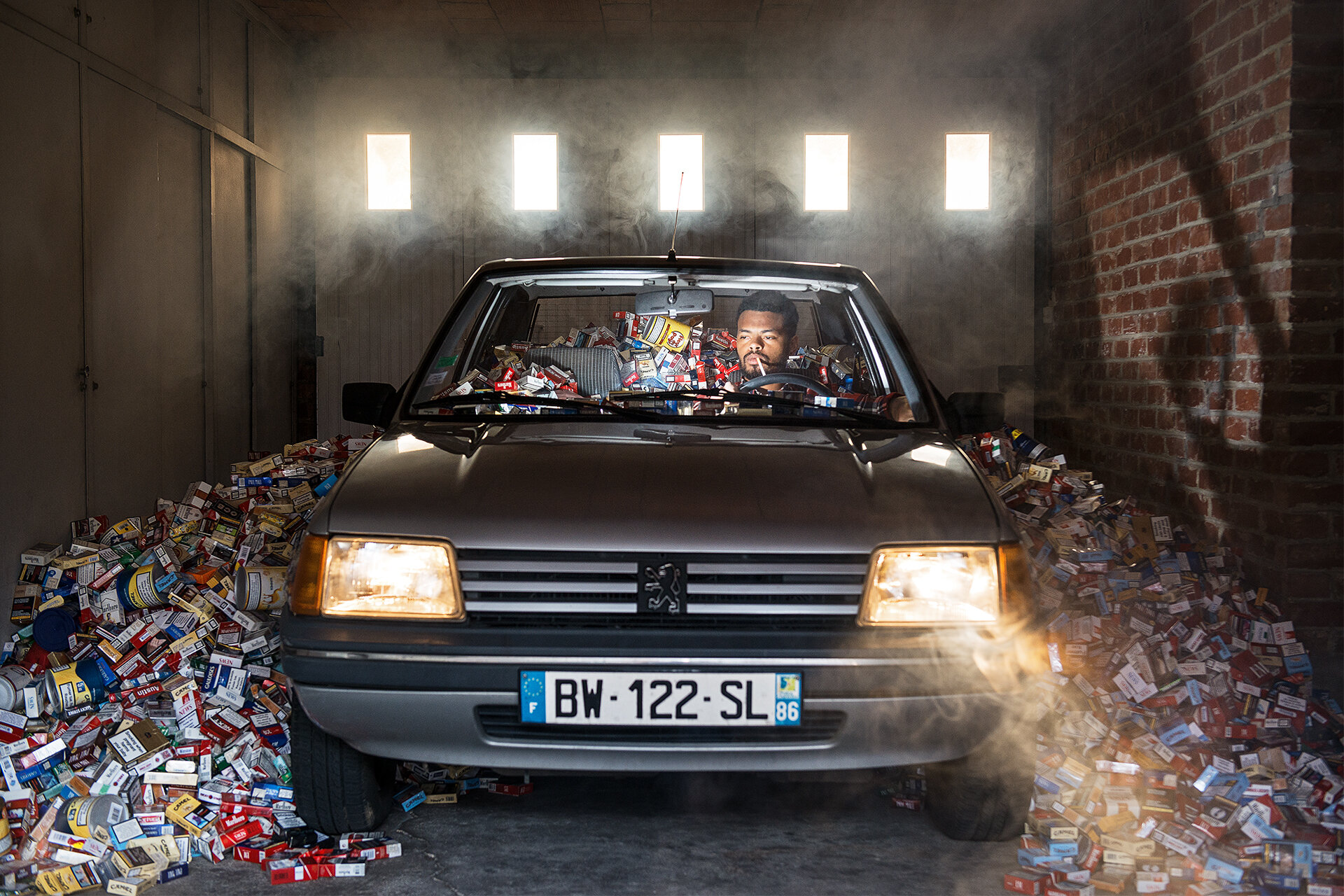 In einer Garage sitzt ein Mann rauchend in seinem Auto. Die Scheinwerfer strahlen in die Kamera. Im Auto und um das Auto herum stapeln sich tausende leerer Zigarettenschachteln.