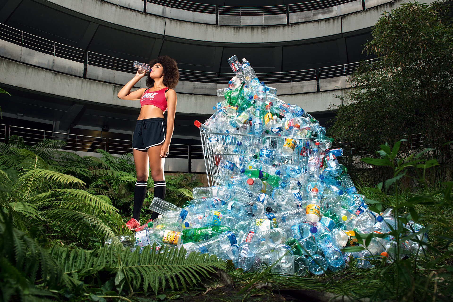Eine junge Frau in Sportkleidung trinkt eine Flasche Wasser, neben ihr ein Warenkorb mit einem riesigen überquellenden Berg an leeren Plastikflaschen. Um sie herum ein altes Gebäude mit mehreren Etagen, welches sich die Natur zurückerobert hat.