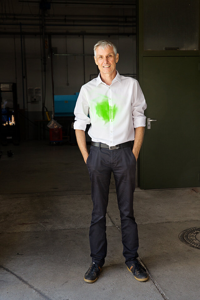 Portrait eines Mannes in weißem Hemd, das hellgrüne Flecke hat in einer dunklen Fabrikhalle.