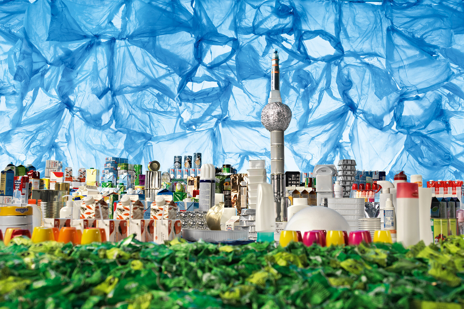 Die Berliner Skyline wird künstlerisch mit buntem Müll inszeniert: Blaue Plastikfolie für den Himmel, Trinkpäckchen, Alufolien und diverser Plastikmüll für die Häuser und den Fernsehturm, grüne Bonbonpapiere für die Grünflächen im Vordergrund.