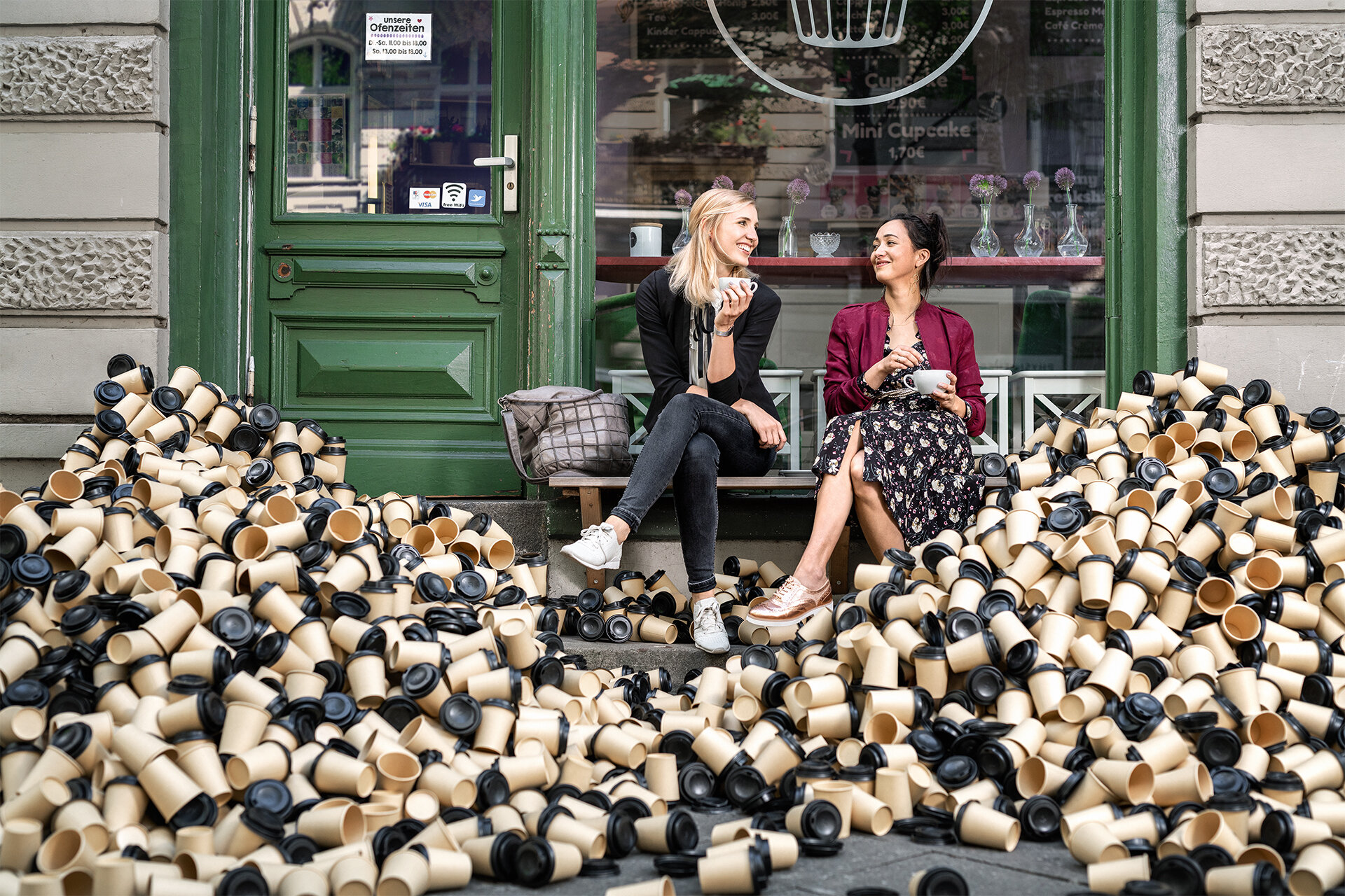 Zwei Frauen sitzen auf einer Bank vor einem Stadtcafé mit ihren Kaffeetassen aus Porzellan. Um sie herum bergweise Wegwerf-Kaffeebecher.