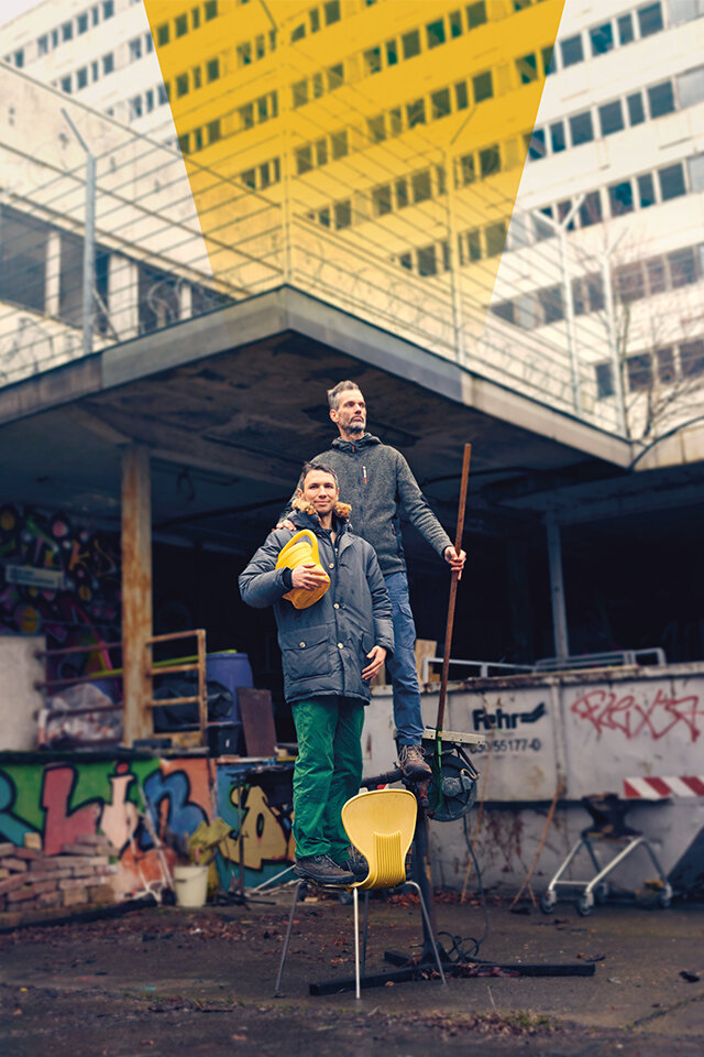 Zwei Männer posieren auf einem Abrissgelände – einer steht mit einer gelben Gießkanne auf einem gelben Stuhl, der andere hält eine Harke. Dieser Mann wird durch ein grafisches, gelbes Element hervorgehoben.