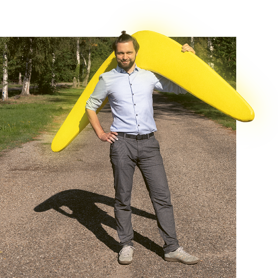 Mann im Businessoutfit trägt menschengroßen, gelben Bumerang über der Schulter und lächelt in die Kamera.