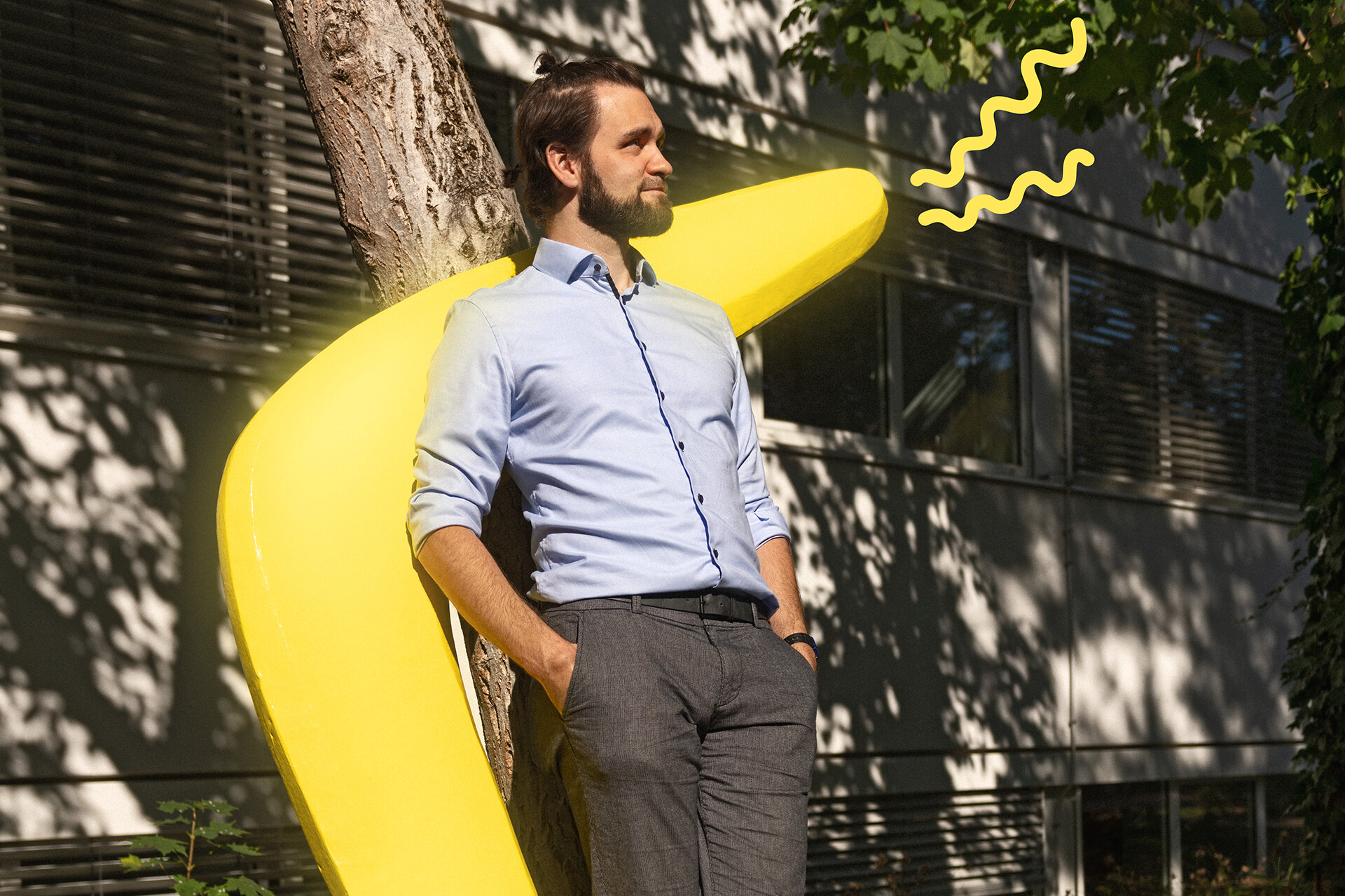 Mann im Businessoutfit lehnt vor einem menschengroßen, gelben Bumerang an einem Baum vor einem Bürogebäude.