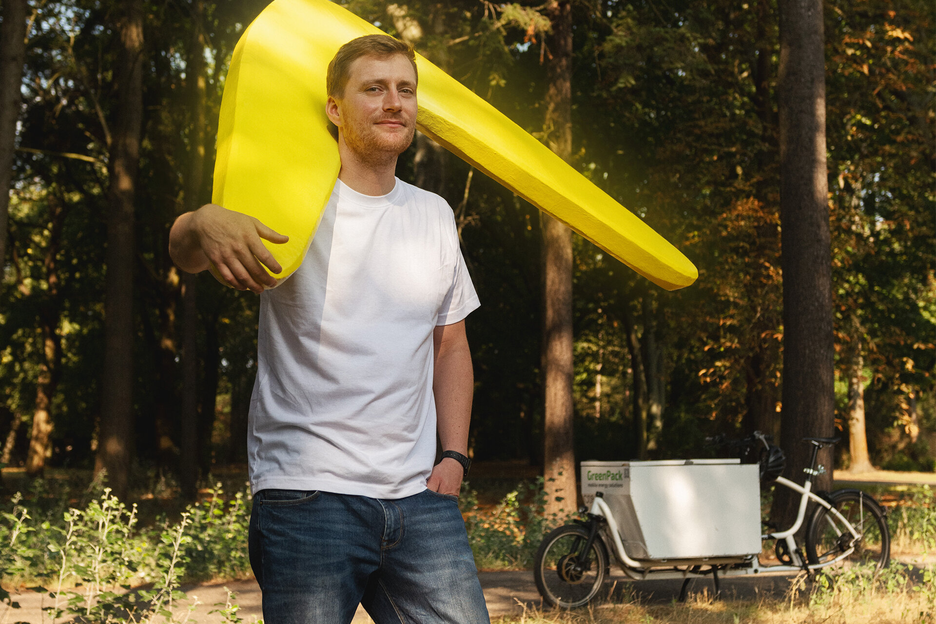 Mann trägt einen menschengroßen, gelben Bumerang über der Schulter. Im Hintergrund ein Lastenfahrrad im Wald.