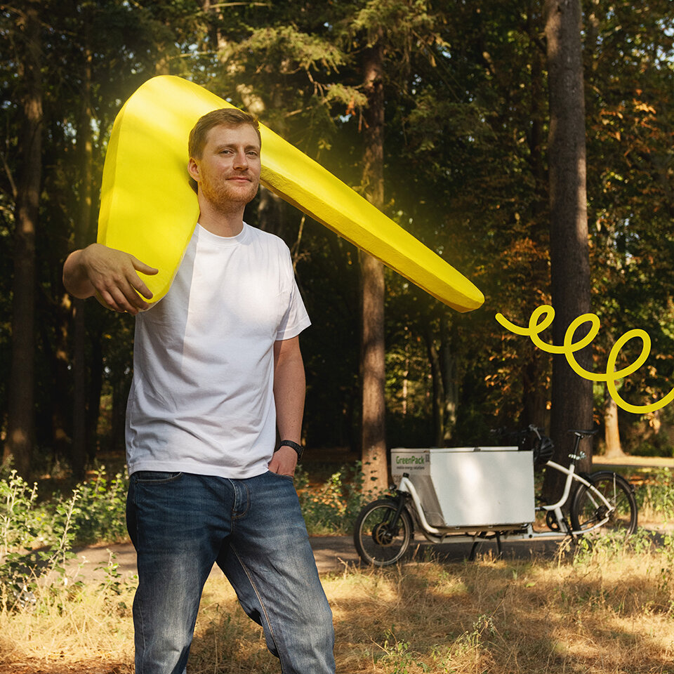 Mann trägt einen menschengroßen, gelben Bumerang über der Schulter. Der Bumerang wird durch ein geschwungenes, grafisches, gelbes Element hervorgehoben. Im Hintergrund ein Lastenfahrrad im Wald.