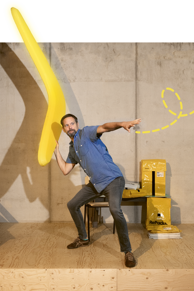 Mann versucht einen menschengroßen, gelben Bumerang zu werfen. Der Bumerang wird durch ein geschwungenes, grafisches, gelbes Element hervorgehoben. Im Hintergrund eine Betonwand hinter Tischen und Stühlen.