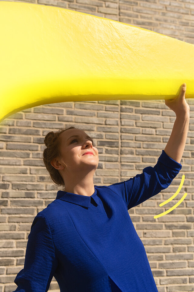 Frau vor einer Backsteinmauer hält einen menschengroßen, gelben Bumerang über ihren Kopf. Der Bumerang wird durch ein geschwungenes, grafisches, gelbes Element hervorgehoben.