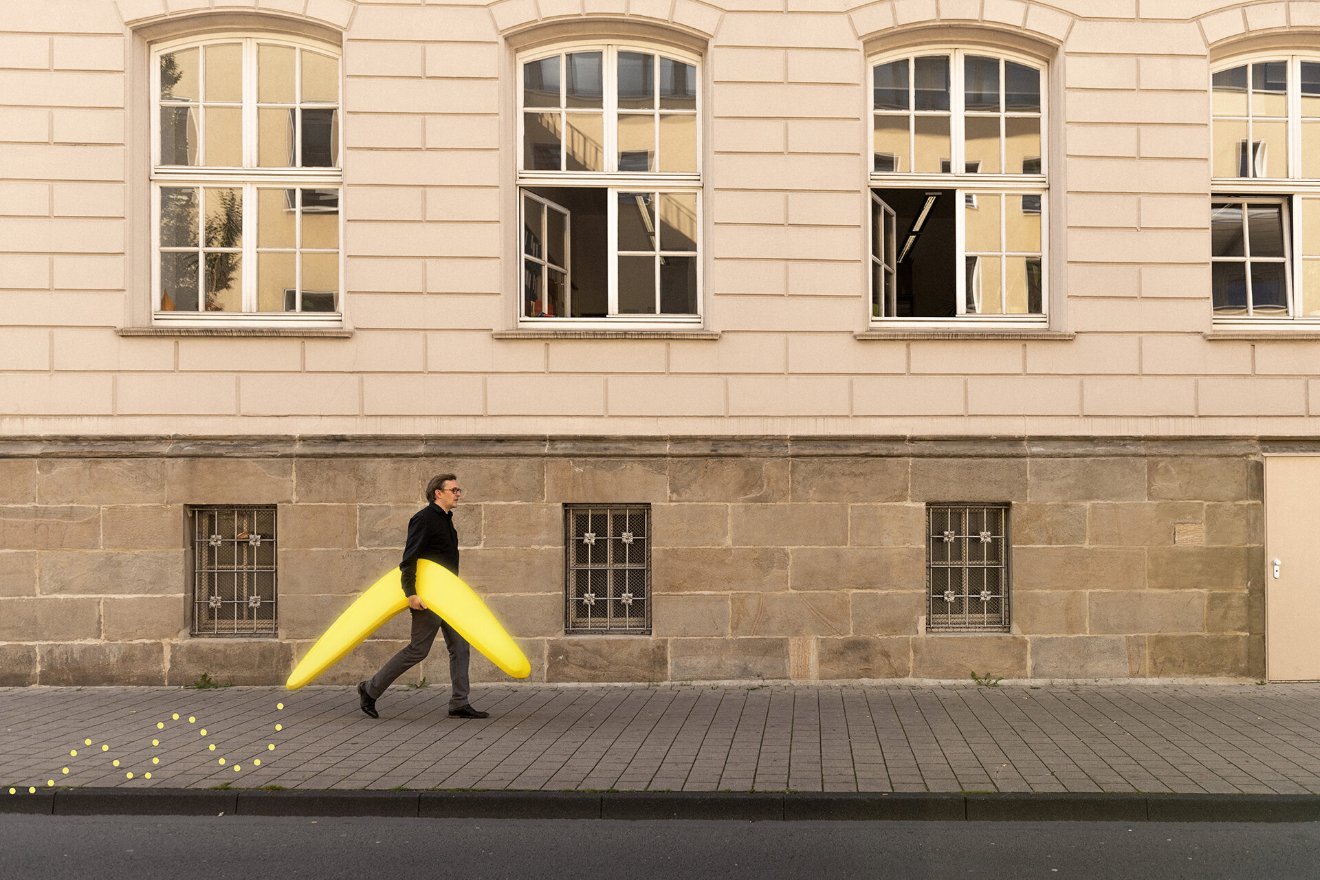 Mann trägt einen menschengroßen, gelben Bumerang unter dem Arm. Der Bumerang wird durch ein gepunktetes, grafisches, gelbes Element hervorgehoben. Im Hintergrund eine Häusermauer.