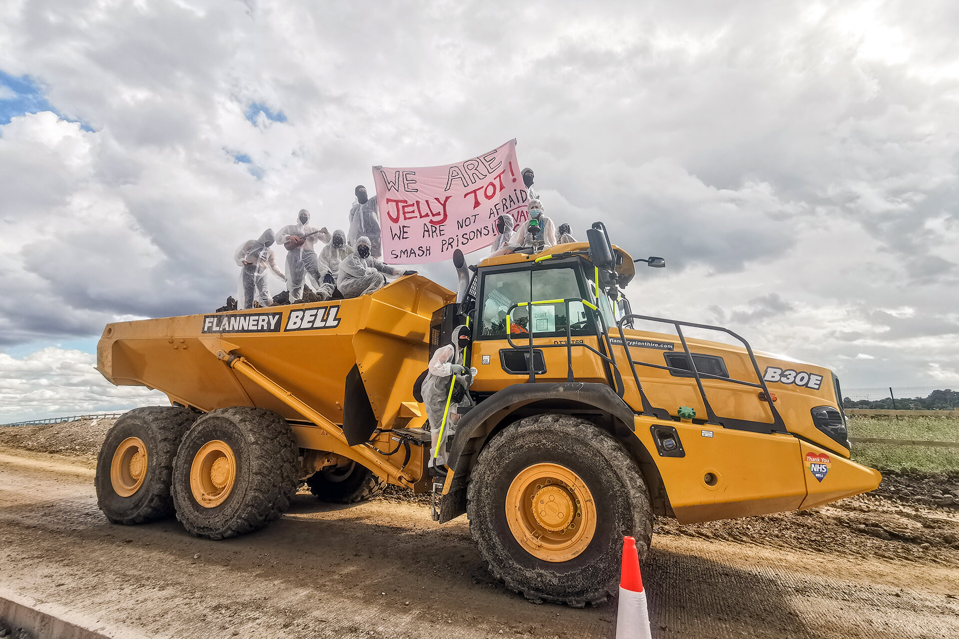 Auf einem Kipplader fahren mehrere in halbtransparenten Overalls gekleidete Waldbesetzer einen Sandweg entlang. Auf ihrem Banner steht: „We are Jelly Tot! We are not afraid. Smash prisons!”