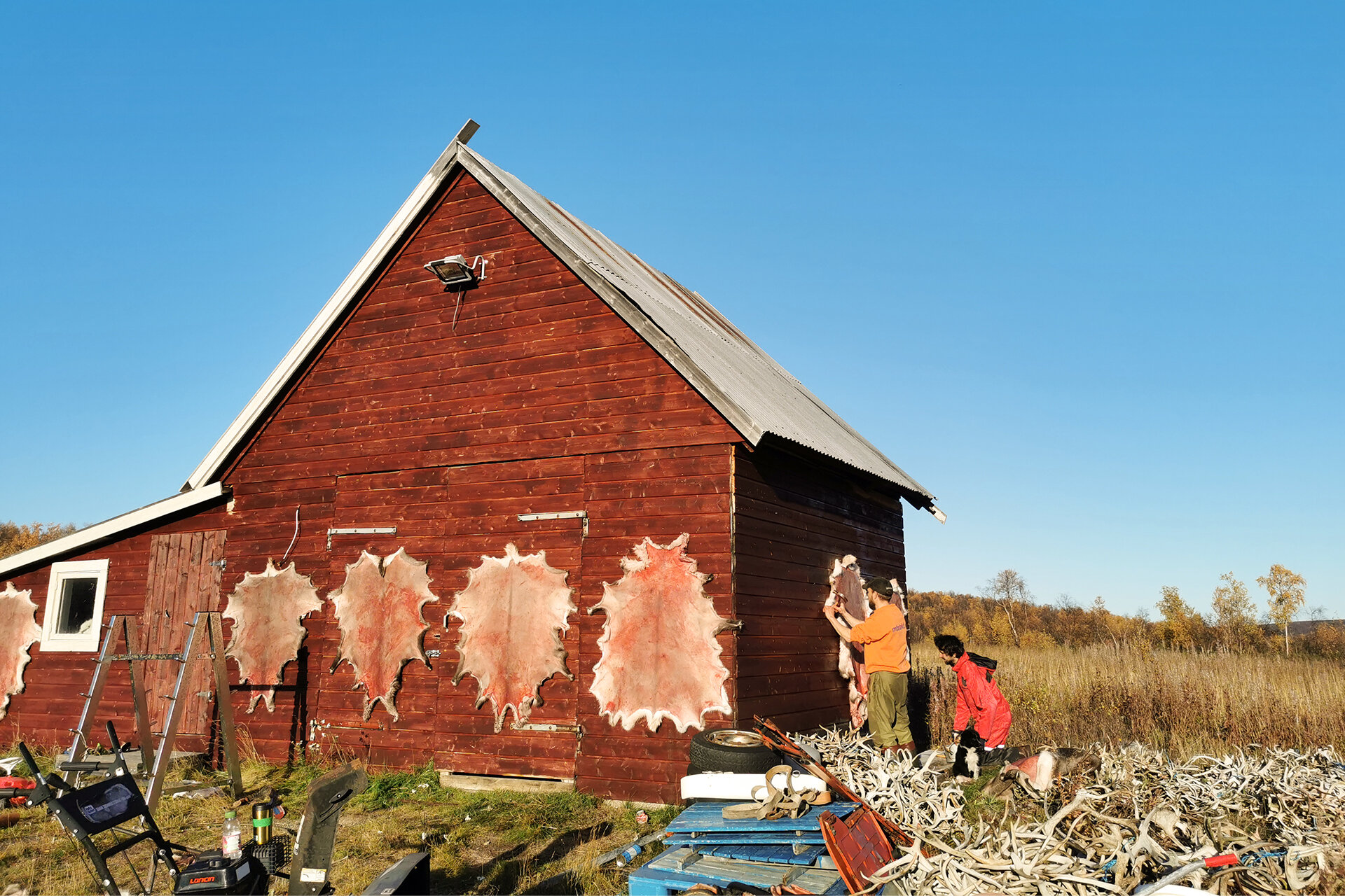 In einer kargen Umgebung steht bei blauem Himmel ein Holzhaus. Um das Haus herum befestigen ein Mann und eine Frau Tierfelle. Im Vordergrund stehen viele Gegenstände herum.