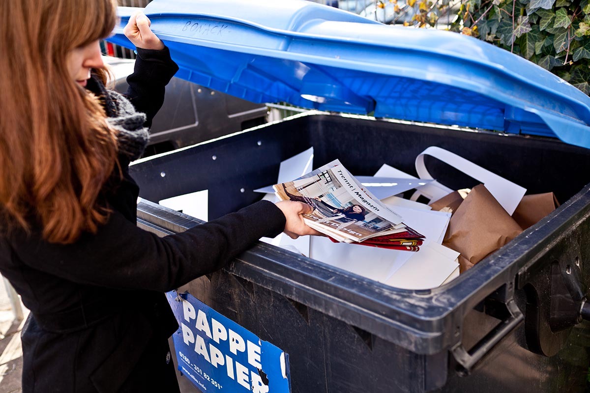 Frau wirft alte Magazine in eine blaue Tonne.