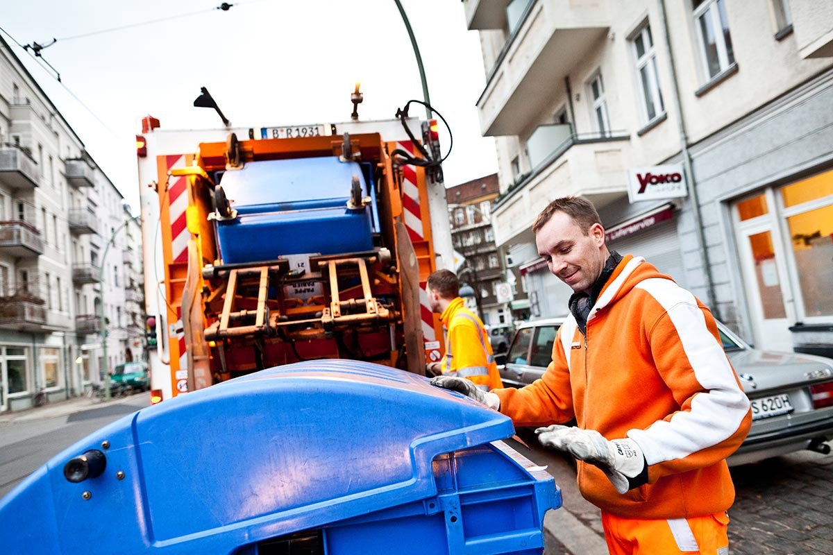 Zwei Männer in orangener Arbeitskleidung stehen an Müllfahrzeug vor blauer Tonne.