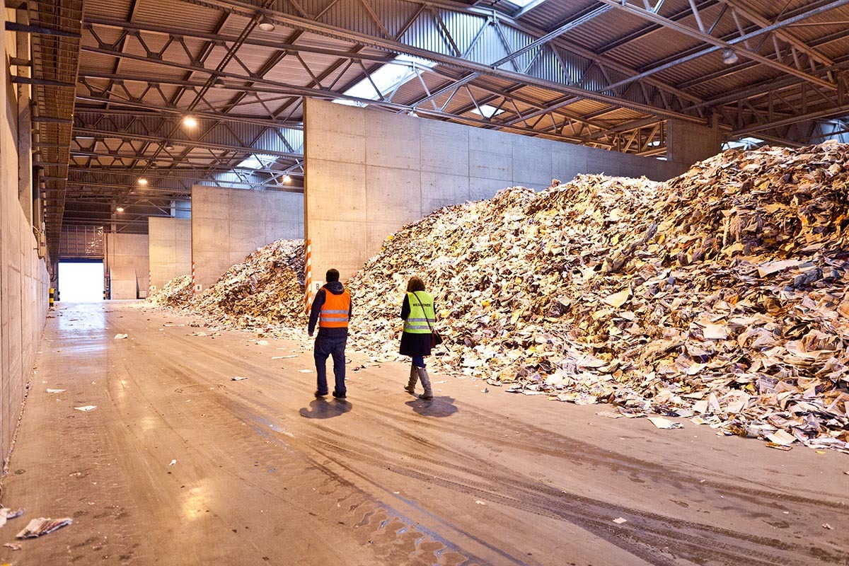 Zwei Menschen laufen durch die Lagerhalle einer Papierfabrik. Vorbei an Altpapier Haufen, die durch hohe Betonwände getrennt sind.