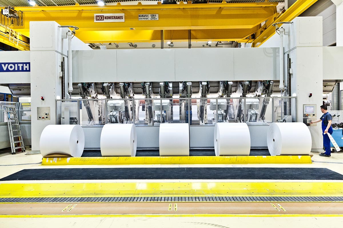 Fünf große Papierrollen in einer Halle. Im Hintergrund eine große Maschine, die von einer Person betätigt wird.