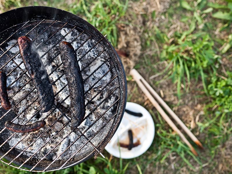 Grill steht auf Wiese. Auf dem Grill liegen verkohlte Würstchen.