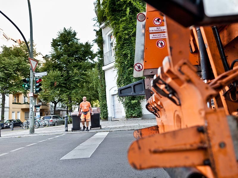 Ein Mann in oranger Arbeitskleidung zieht zwei schwarze Mülltonnen zum Müllfahrzeug über die Straße.