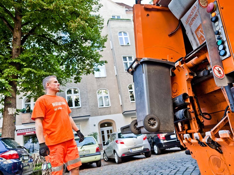 Ein Mann in oranger Arbeitskleidung sieht zu, wie eine Mülltonne vom Müllfahrzeug zur Entleerung angehoben wird.