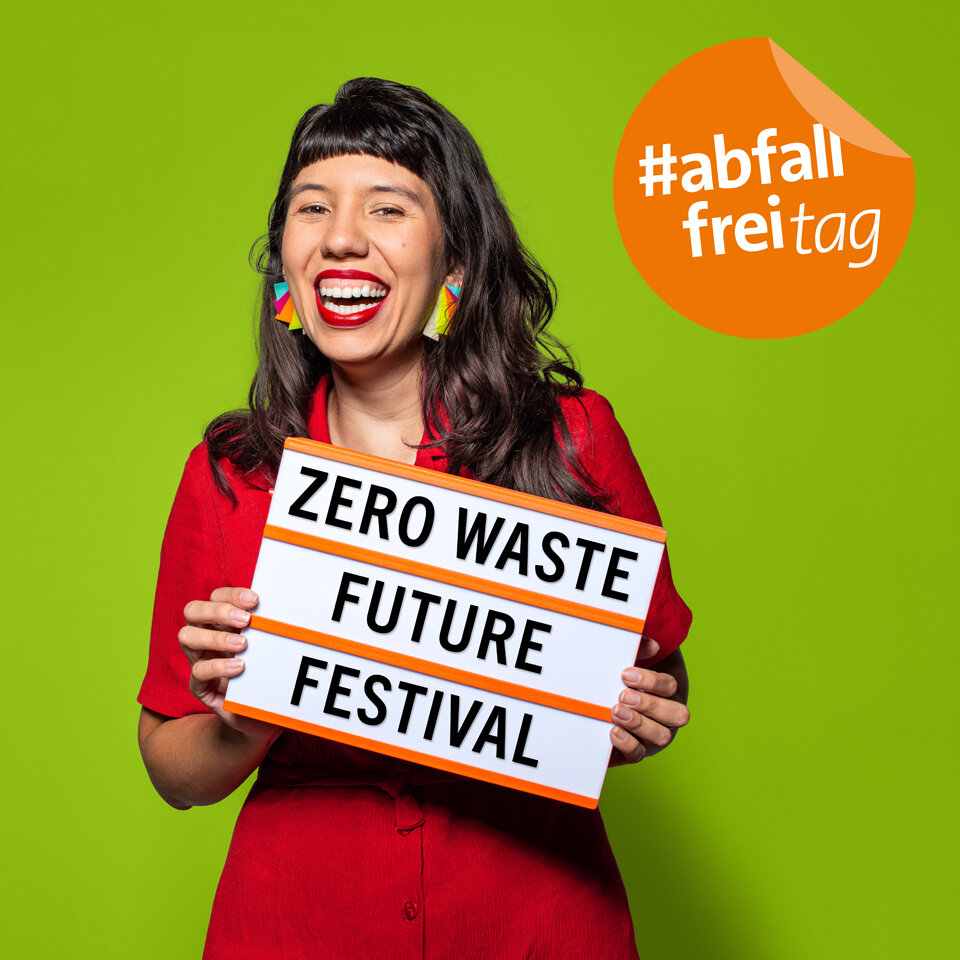 Frau in rotem Kleid steht vor grünem Hintergrund und hält eine Leuchtbox mit dem Text Zero Waste Fututre Festival